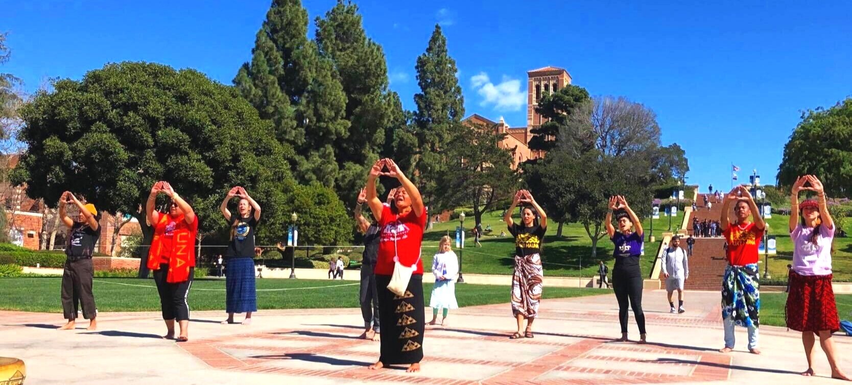 Mauna Kea Protectors at the base of a series of steps chanting and praying for the rejection to TMT. The background features a blue sky and green trees and some passerbys on the lawn.